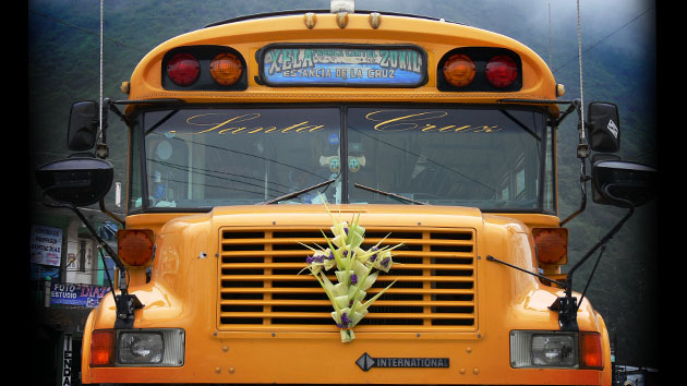 A yellow school bus is adorned at its front with a cross made of flowers and broad folded leaves