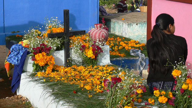 A white tomb is adorned with marigolds, greenery, flowers, vases, and a large black cross