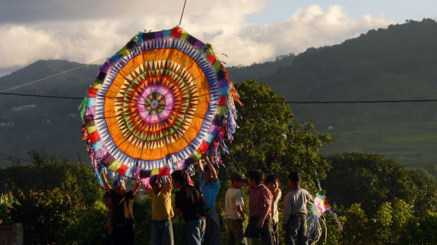 Outdoors in sunlight, several people hold a large circular kite with colorful patterns aloft.