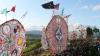 Circular, colorful patterned kites as tall as trees stand at the edge of a field with people below.